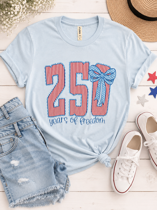 Light blue t-shirt with '250 years of freedom' design, denim shorts, white sneakers, and a hat on a wooden surface.