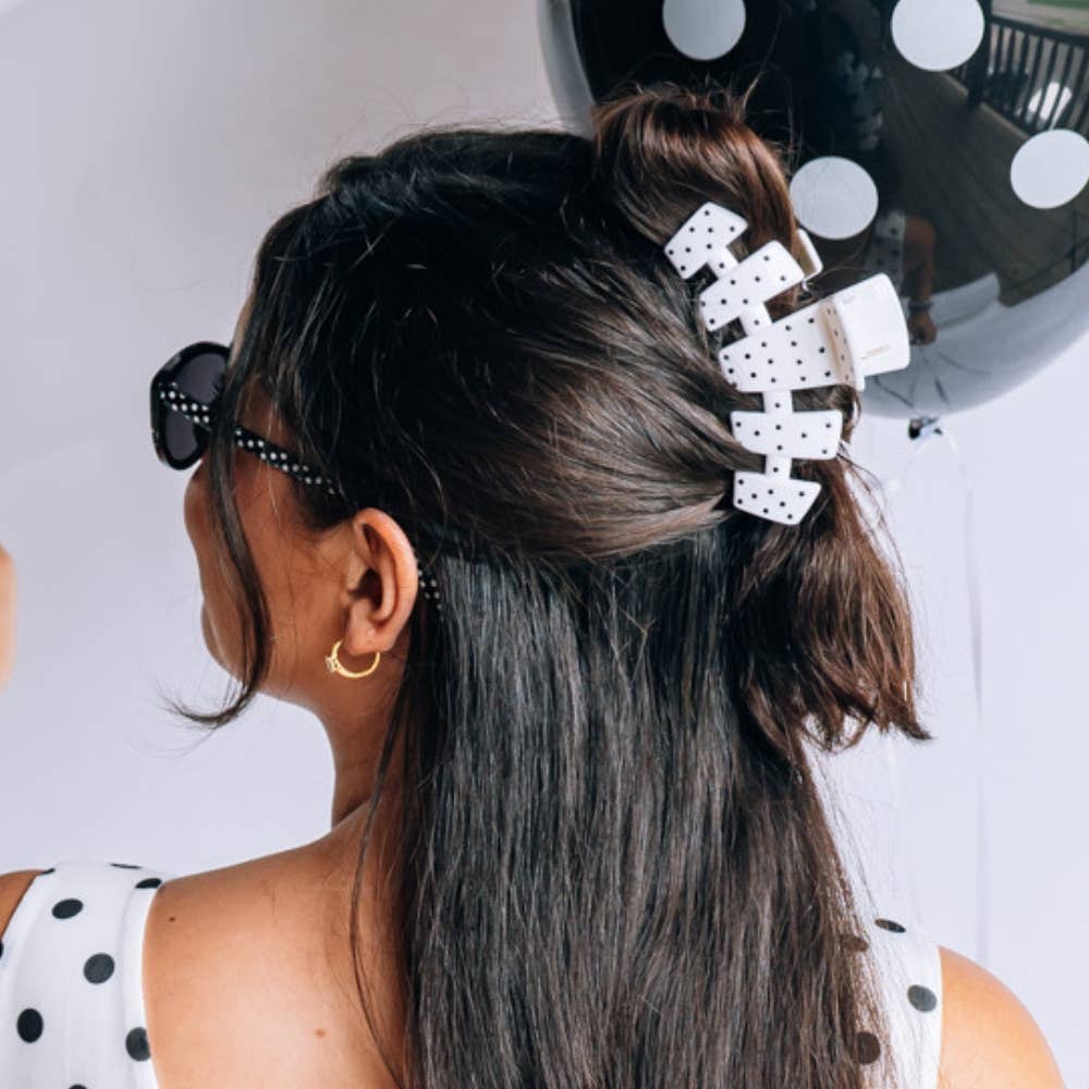 A woman shown from the back wearing a white hair claw clip with black polka dots, holding her dark hair in a half-up style. She is also wearing a matching white top with black polka dots and black sunglasses.