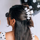 A woman shown from the back wearing a white hair claw clip with black polka dots, holding her dark hair in a half-up style. She is also wearing a matching white top with black polka dots and black sunglasses.