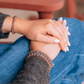 Close-up of a person's hands resting on their lap, wearing three brown bracelets, one on each wrist, with blue jeans and a dark gray sweater visible.