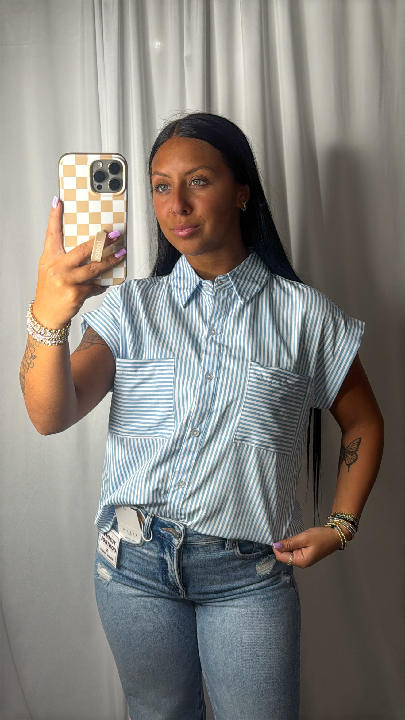 A woman taking a mirror selfie wearing a light blue and white vertically striped short-sleeve button-up shirt and blue jeans, holding a phone with a checkered case against a gray curtain backdrop.
