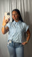 A woman taking a mirror selfie wearing a light blue and white vertically striped short-sleeve button-up shirt and blue jeans, holding a phone with a checkered case against a gray curtain backdrop.