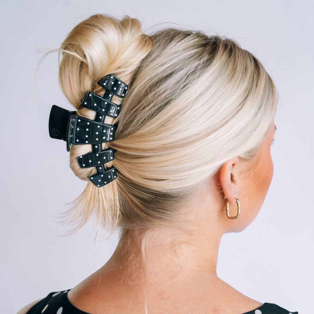 A blonde woman shown from the back wearing a black claw hair clip with white polka dots, holding her hair in a bun. She is wearing a black and white polka dot top and a gold hoop earring against a light gray background.