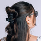 A woman shown from the back and side wearing a black hair claw clip with white polka dots, holding her dark brown hair in a low bun. She is wearing black sunglasses with white polka dot frames and a white top with black polka dots, against a light gray background.