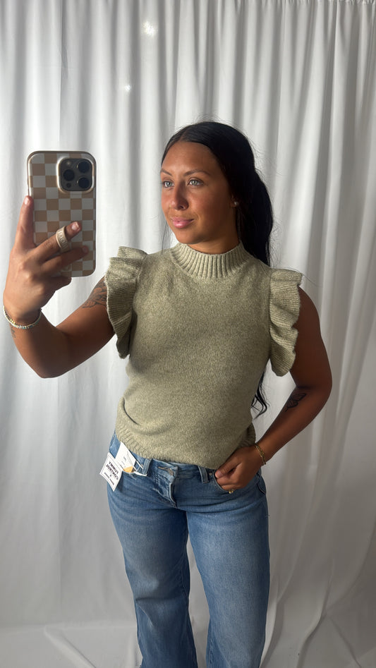 A woman takes a mirror selfie wearing a light sage green knit sweater with ruffled shoulder details and blue jeans, standing in front of a white curtain backdrop.