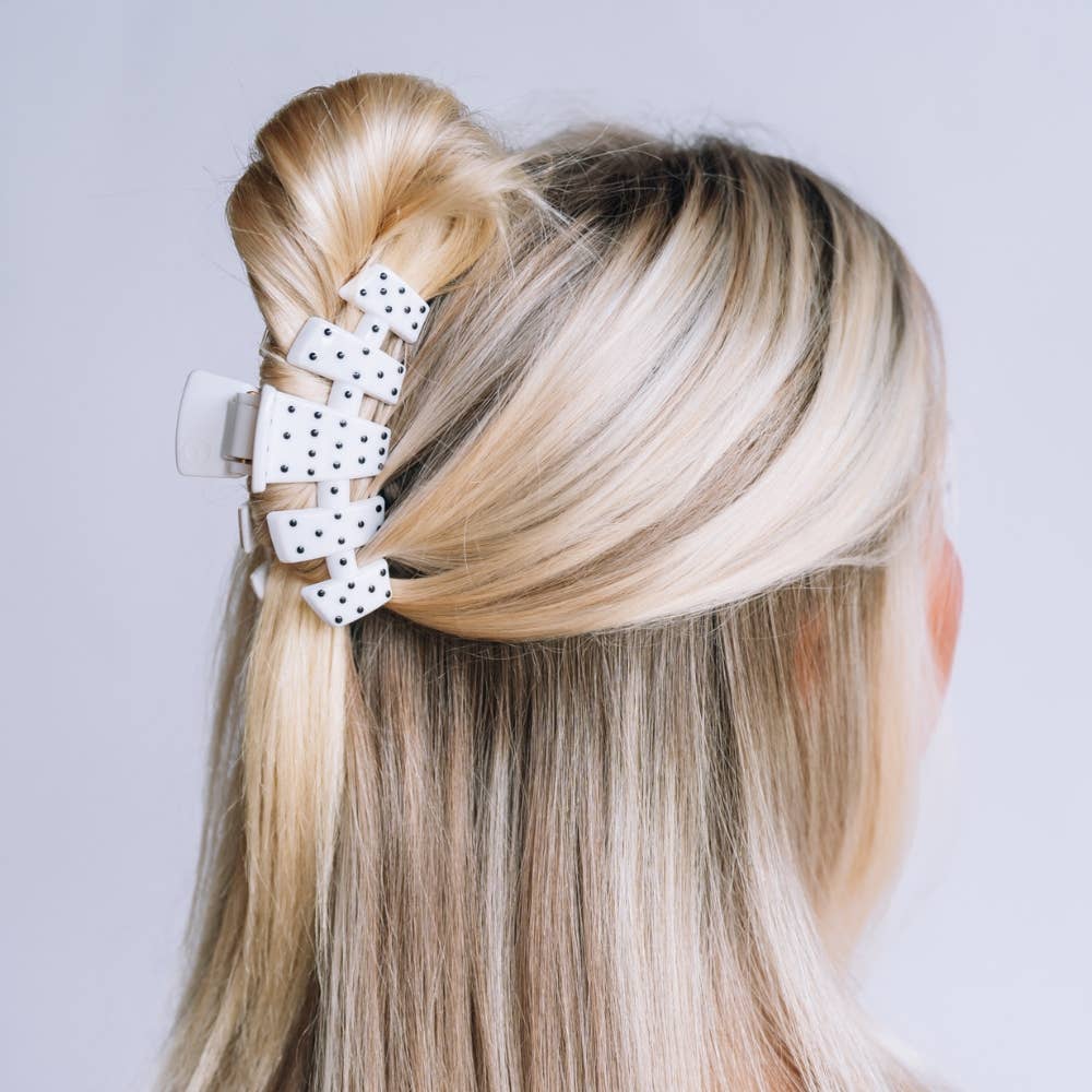 A blonde woman shown from the back wearing a white hair claw clip with black polka dots, holding her hair in a half-up style against a light gray background.