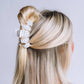 A blonde woman shown from the back wearing a white hair claw clip with black polka dots, holding her hair in a half-up style against a light gray background.