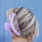 Person with blonde hair tied back with a purple hair clip against a blue background