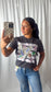 A woman taking a mirror selfie wearing a dark gray graphic t-shirt with a white dove design and blue jeans, holding a checkered phone case against a white curtain backdrop.
