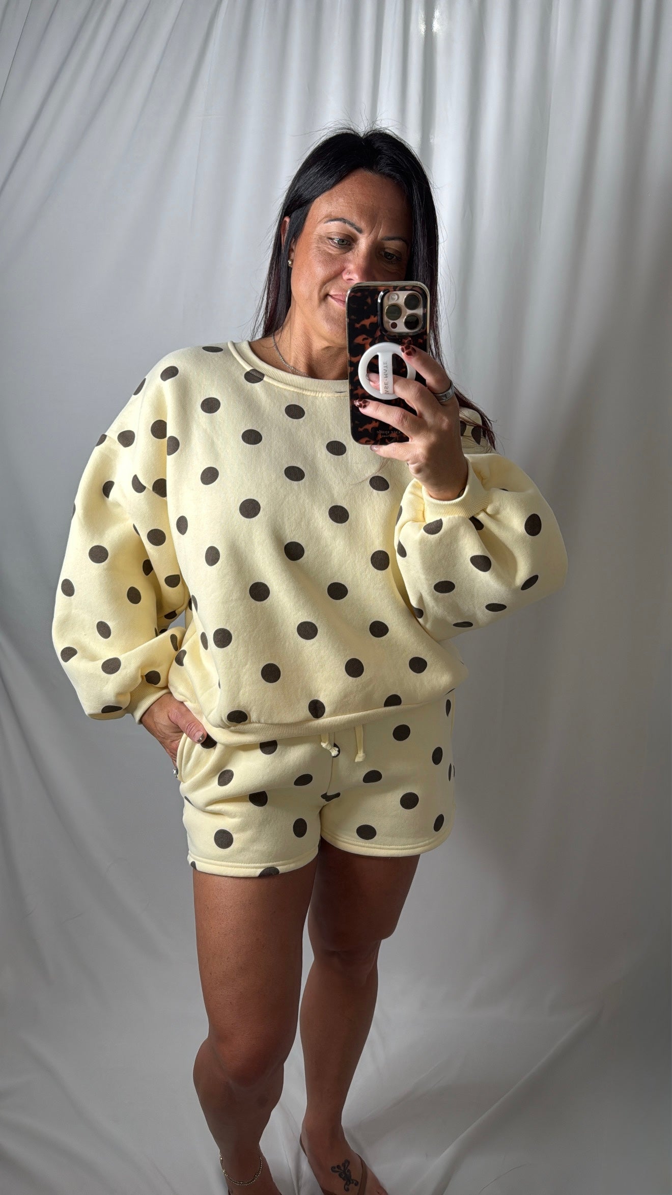 A woman takes a mirror selfie wearing a butter yellow-colored two-piece loungewear set with brown polka dots, with a white background, travel, set