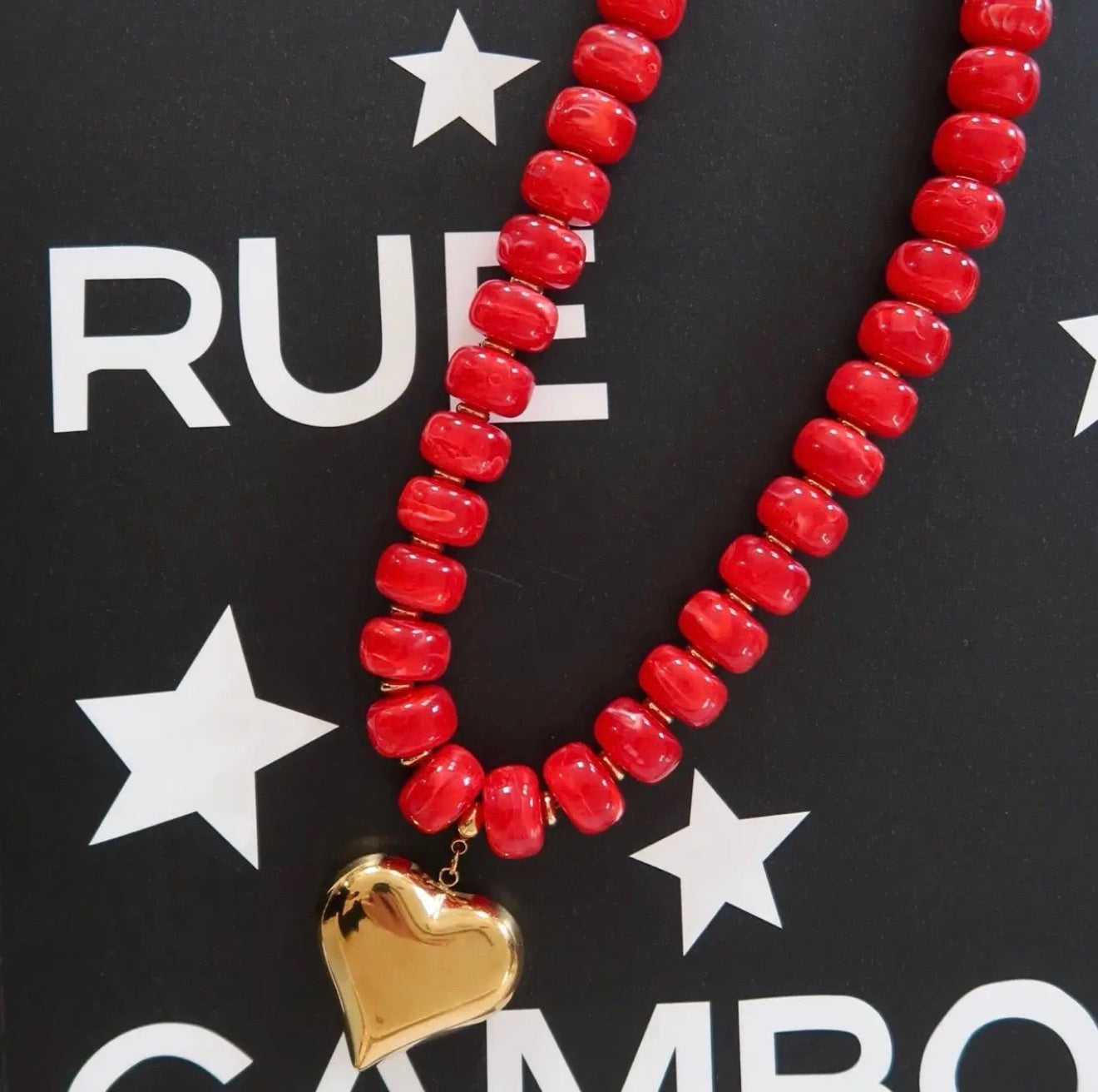 A red beaded necklace with a shiny gold heart pendant, displayed on a black background with white star and text graphics.