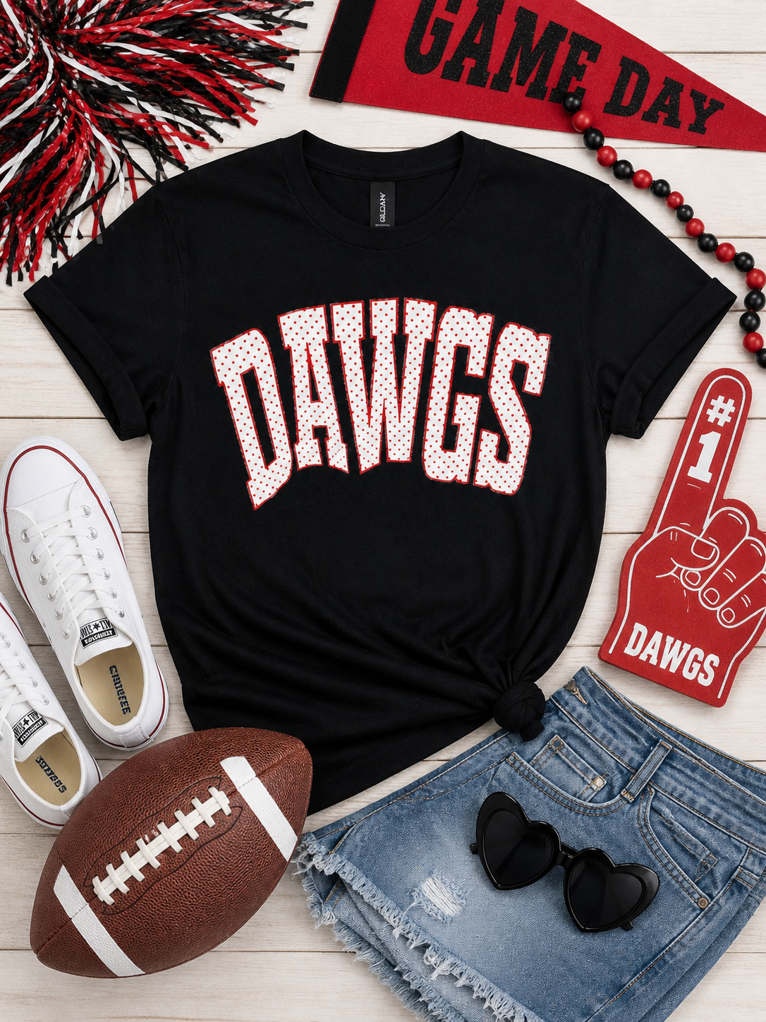 A black short-sleeve graphic T-shirt laid flat on a light wood surface, styled with a football, shorts, sunglasses, sneakers, a pennant, beads, and a foam finger around it. The shirt has large arched 'DAWGS' lettering across the chest in white with a red dotted outline.