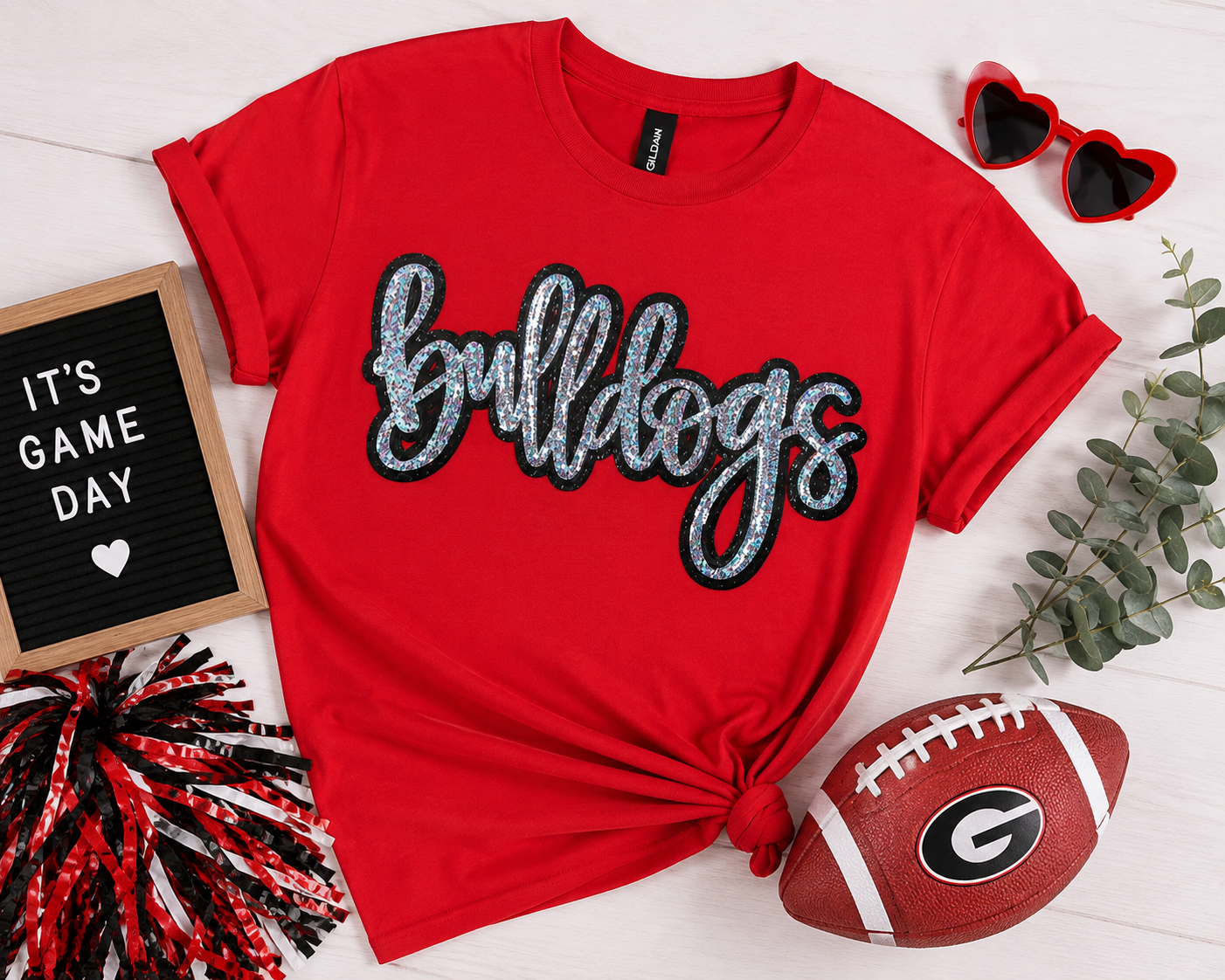 A red short-sleeve T-shirt with 'bulldogs' in glittery silver script outlined in black across the chest, styled in a flat lay with a tied hem. The shirt is surrounded by football-themed props including a brown football with a Georgia 'G' logo, red heart-shaped sunglasses, a letter board reading 'IT'S GAME DAY', and greenery on a white wood background.