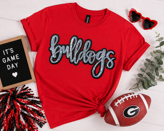 A red short-sleeve T-shirt with 'bulldogs' in glittery silver script outlined in black across the chest, styled in a flat lay with a tied hem. The shirt is surrounded by football-themed props including a brown football with a Georgia 'G' logo, red heart-shaped sunglasses, a letter board reading 'IT'S GAME DAY', and greenery on a white wood background.