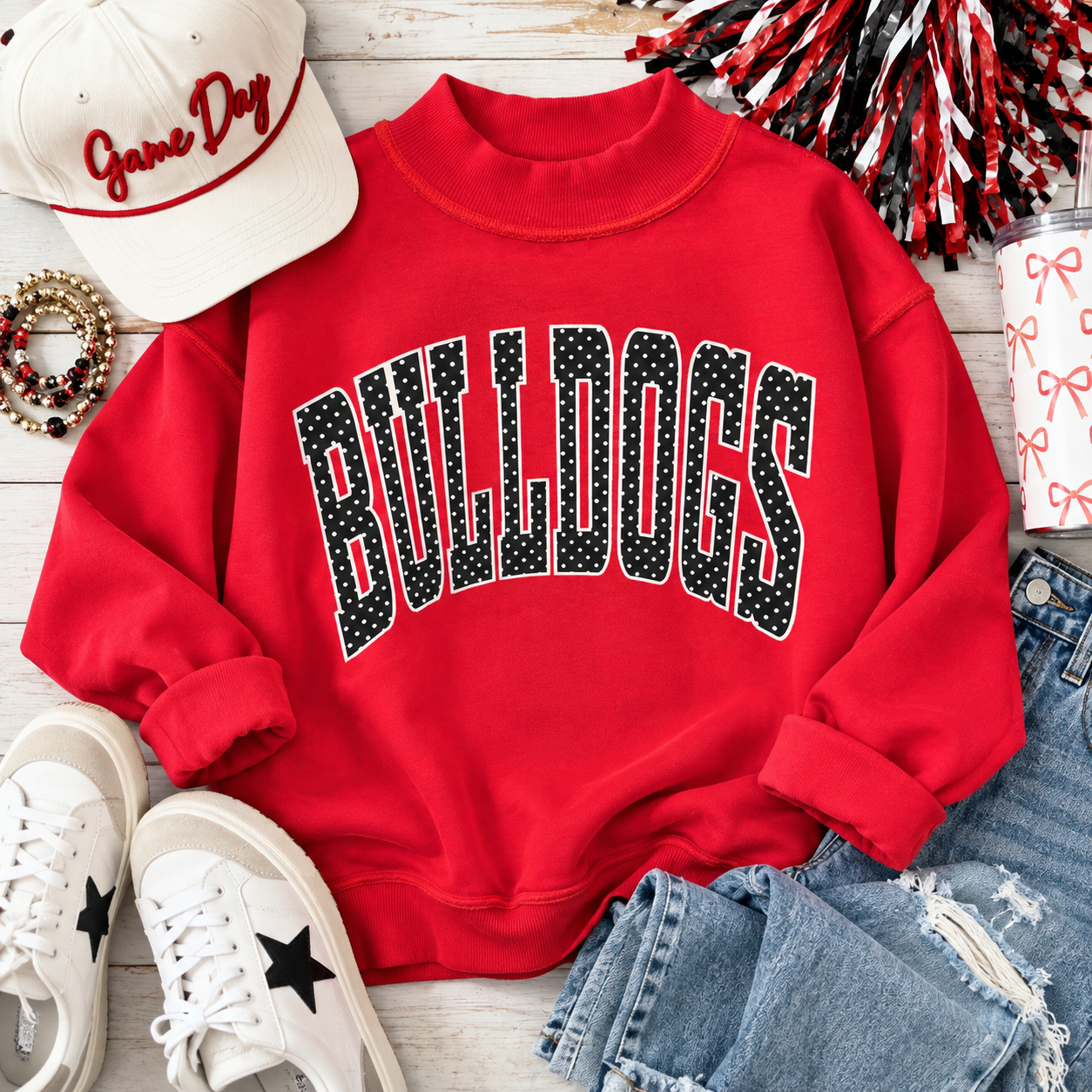 A red crewneck sweatshirt with 'BULLDOGS' in a black-and-white polka dot arched font across the chest, styled in a flat lay with a white cap, white sneakers, blue jeans, and red-and-white pom-poms on a whitewashed wood background.