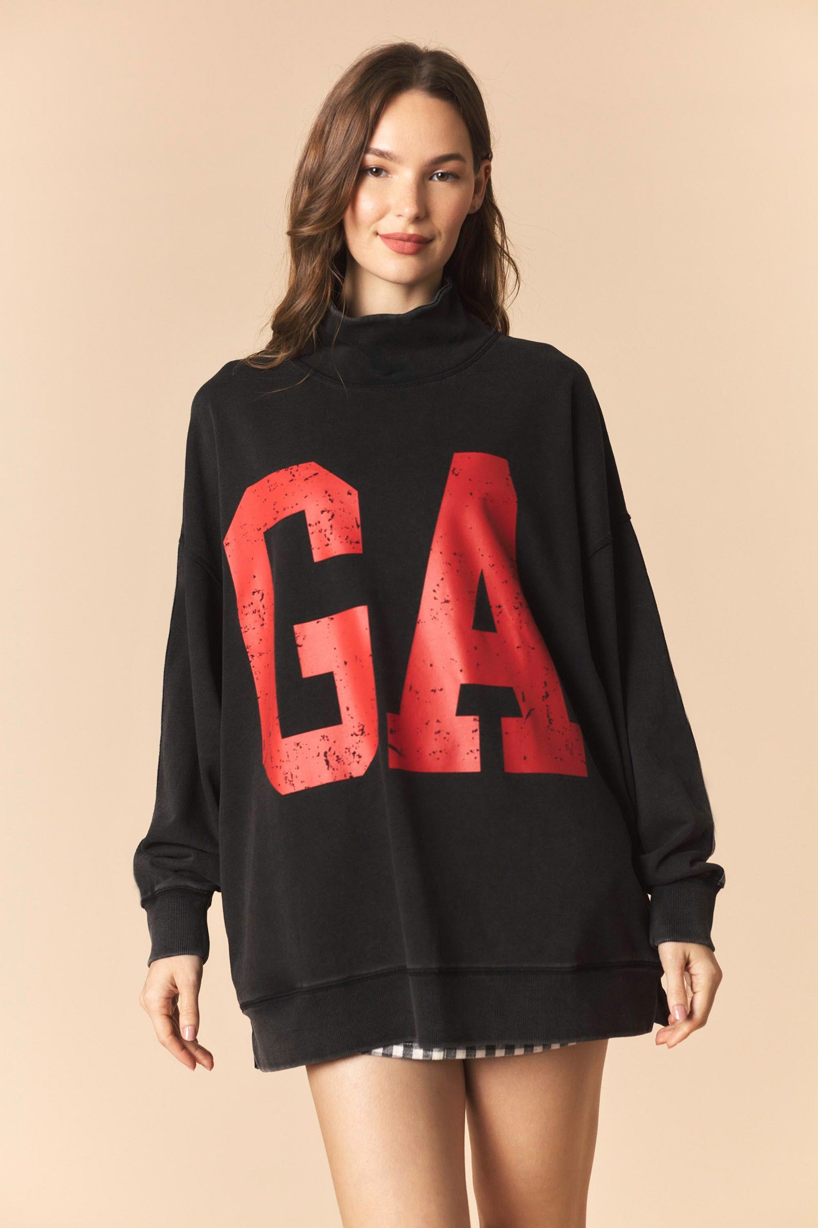 A woman modeling an oversized black sweatshirt with a large distressed red 'GA' graphic on the front, set against a solid beige background.