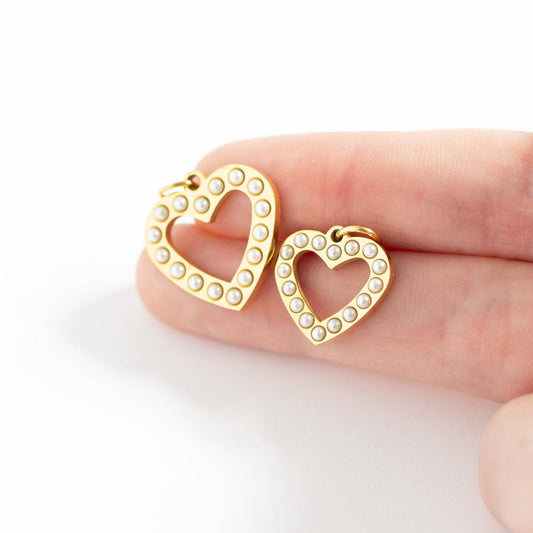 Gold heart-shaped earrings with pearl accents held between fingers on a white background