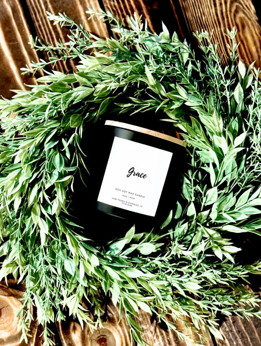 Candle labeled 'Grace' surrounded by green leaves on a wooden surface