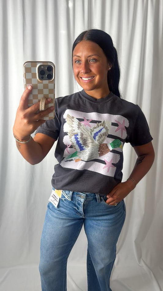 A woman taking a mirror selfie wearing a dark gray graphic t-shirt with a white dove and pink star design, paired with blue jeans, against a white curtain backdrop.