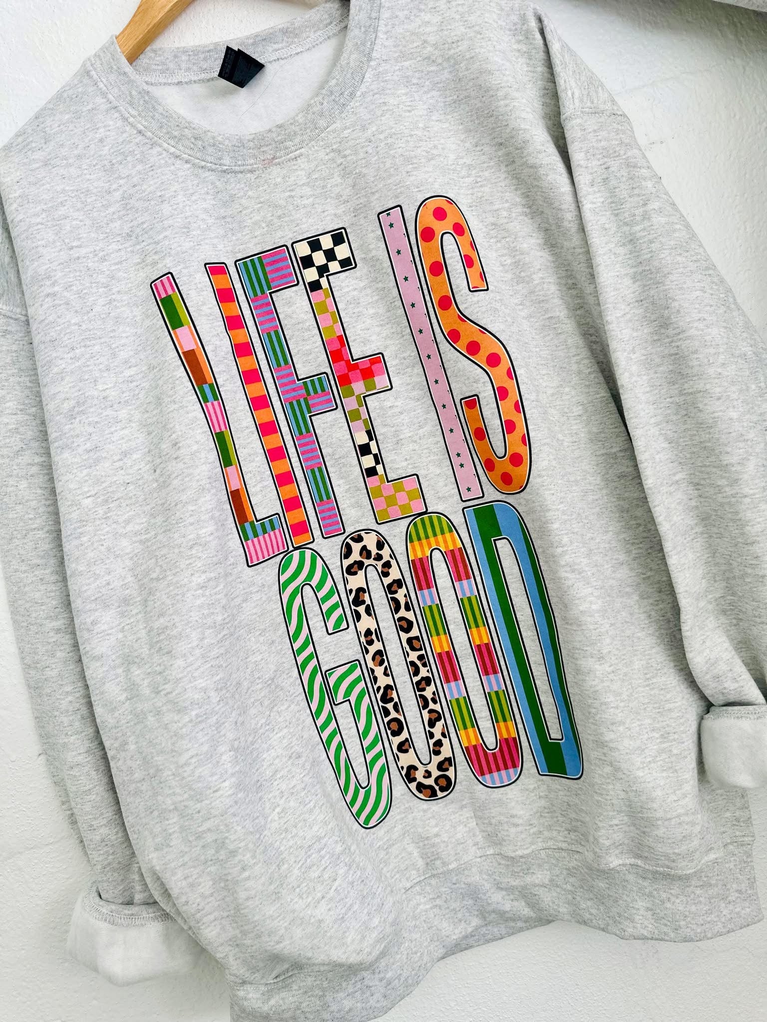 Gray sweatshirt with 'LIFE IS GOOD' text in colorful patterns on a white background
