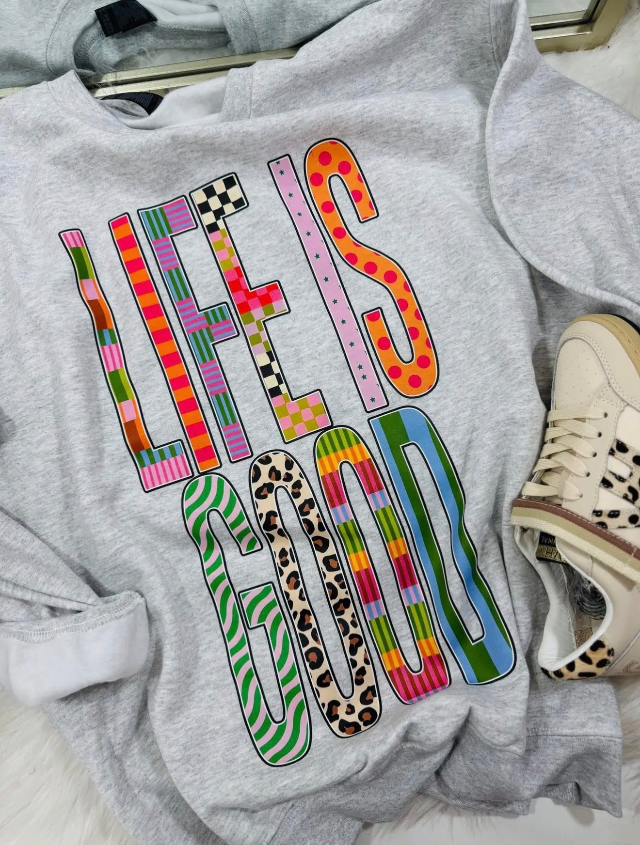 Gray sweatshirt with 'Life is Good' text in colorful patterns on a white surface.