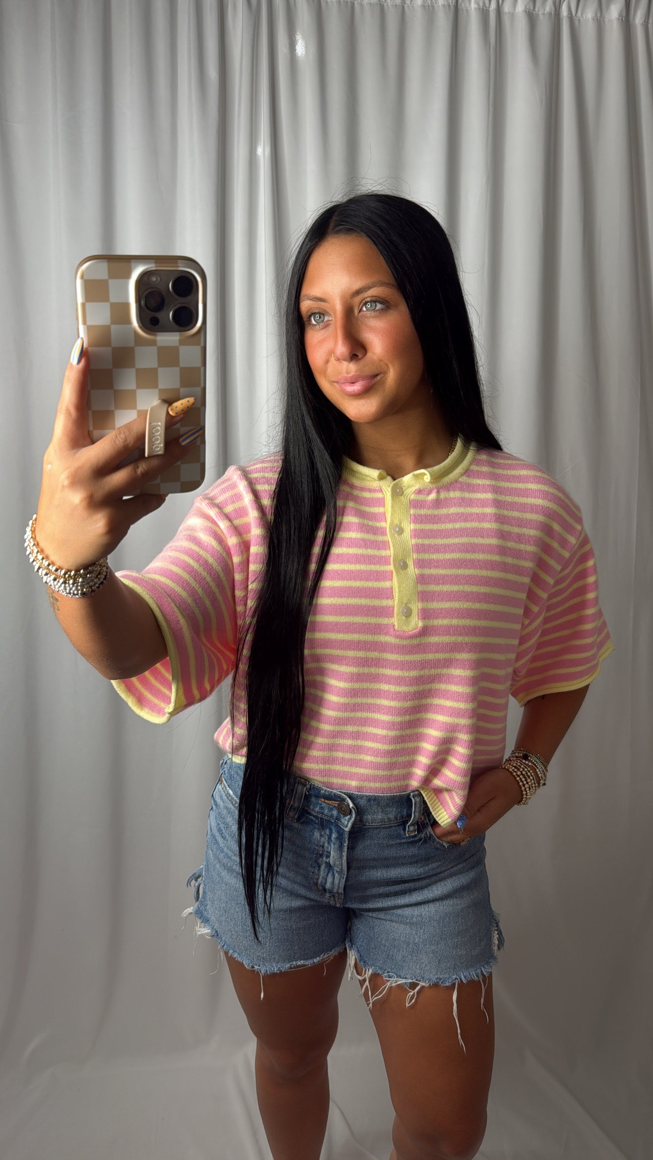 A woman taking a mirror selfie wearing a pink and white striped short-sleeve henley shirt with yellow trim, paired with denim shorts, against a white curtain backdrop.