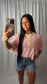 A woman taking a mirror selfie wearing a pink and white striped short-sleeve henley shirt with yellow trim, paired with denim shorts, against a white curtain backdrop.
