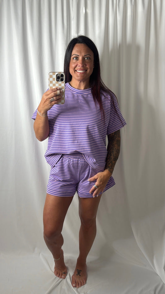 A woman takes a mirror selfie wearing a matching purple and pink striped lounge set consisting of a short-sleeve t-shirt and shorts, standing against a white draped fabric backdrop.