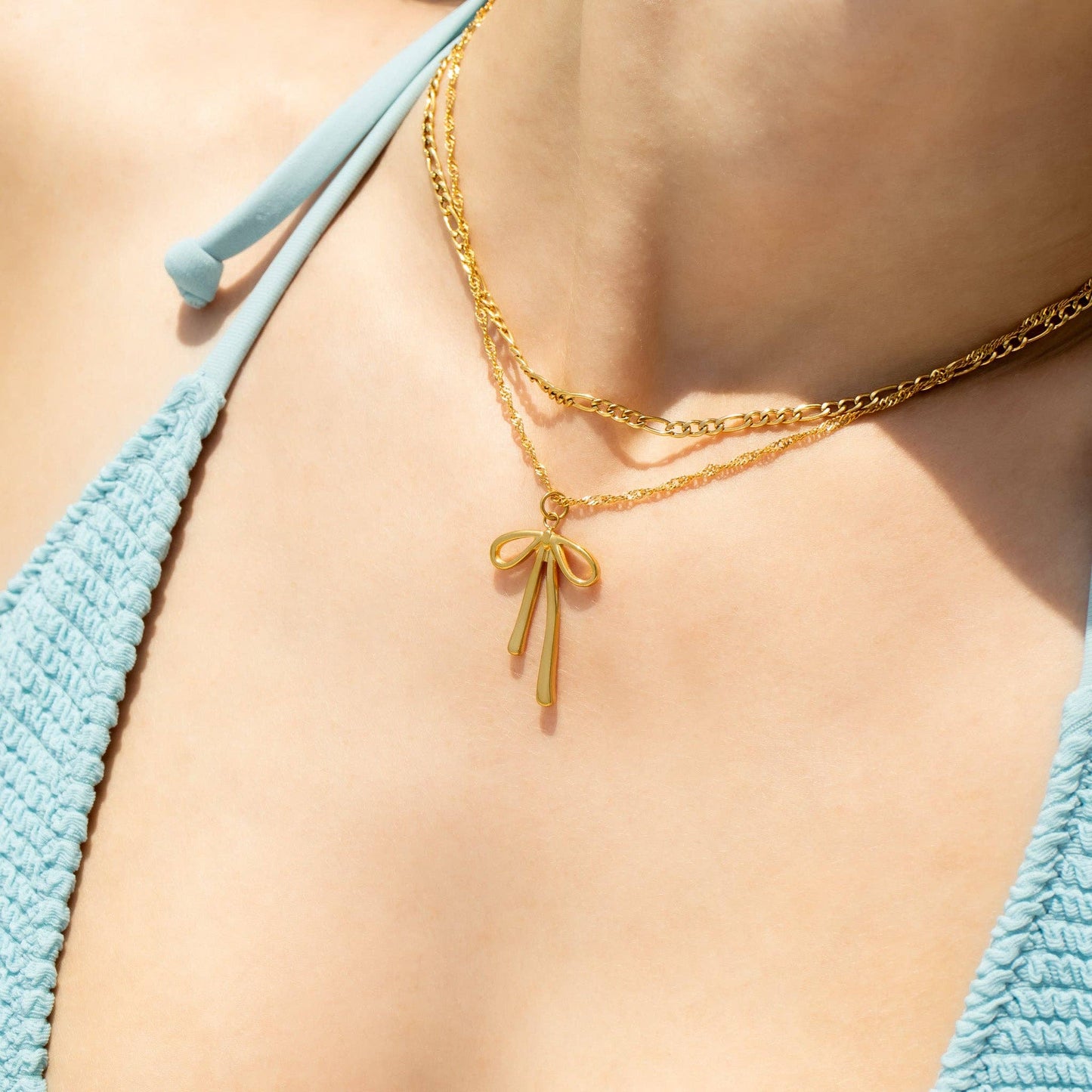 Gold necklace with a bow pendant worn by a person in a blue bikini.