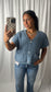 A woman takes a mirror selfie wearing a light blue textured short-sleeve cardigan with front buttons and pockets, paired with blue jeans, against a white curtain backdrop.