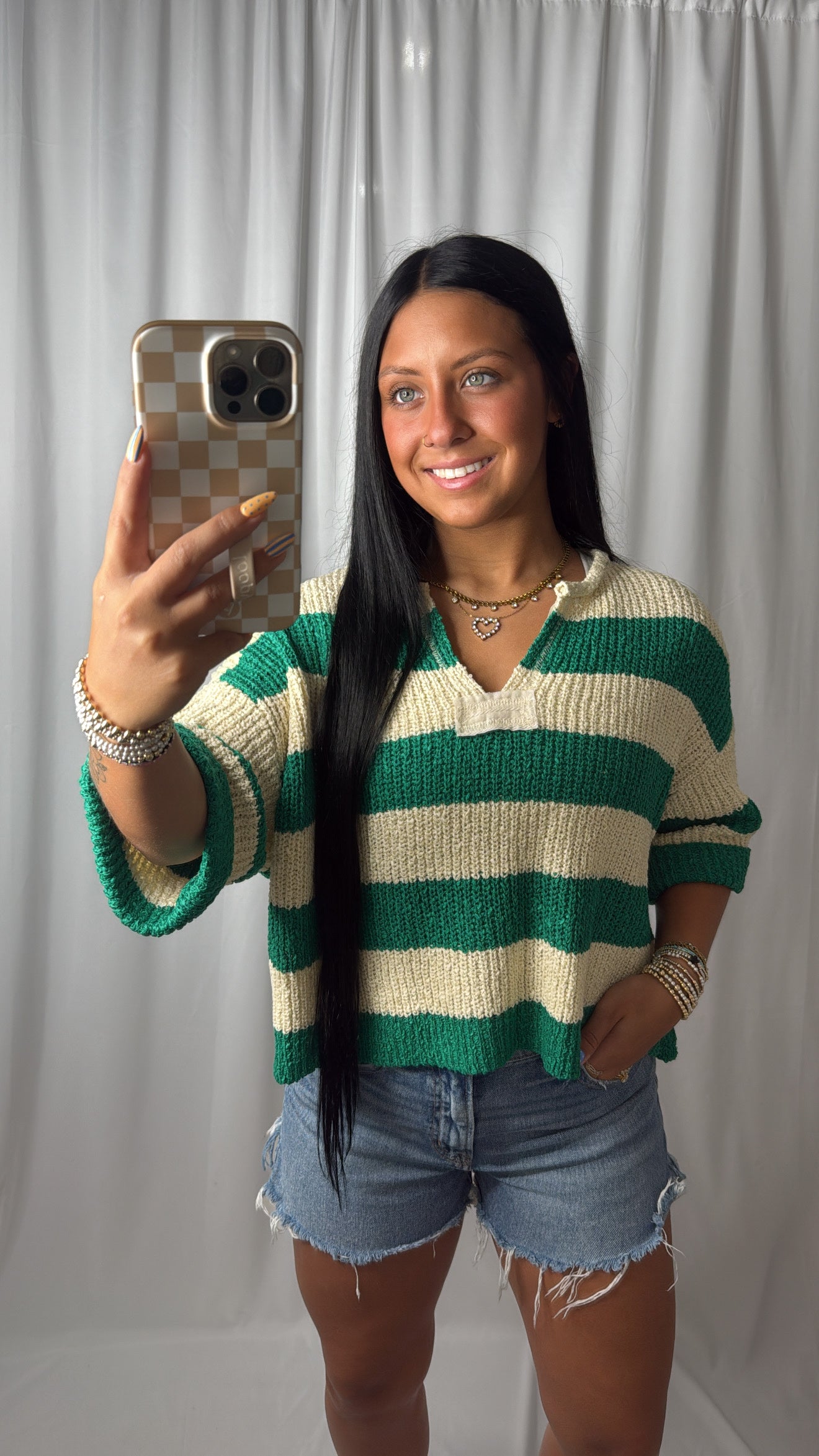 A woman taking a mirror selfie wearing a green and cream striped knit sweater, denim shorts, and jewelry against a white curtain backdrop.