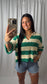 A woman taking a mirror selfie wearing a green and cream striped knit sweater, denim shorts, and jewelry against a white curtain backdrop.