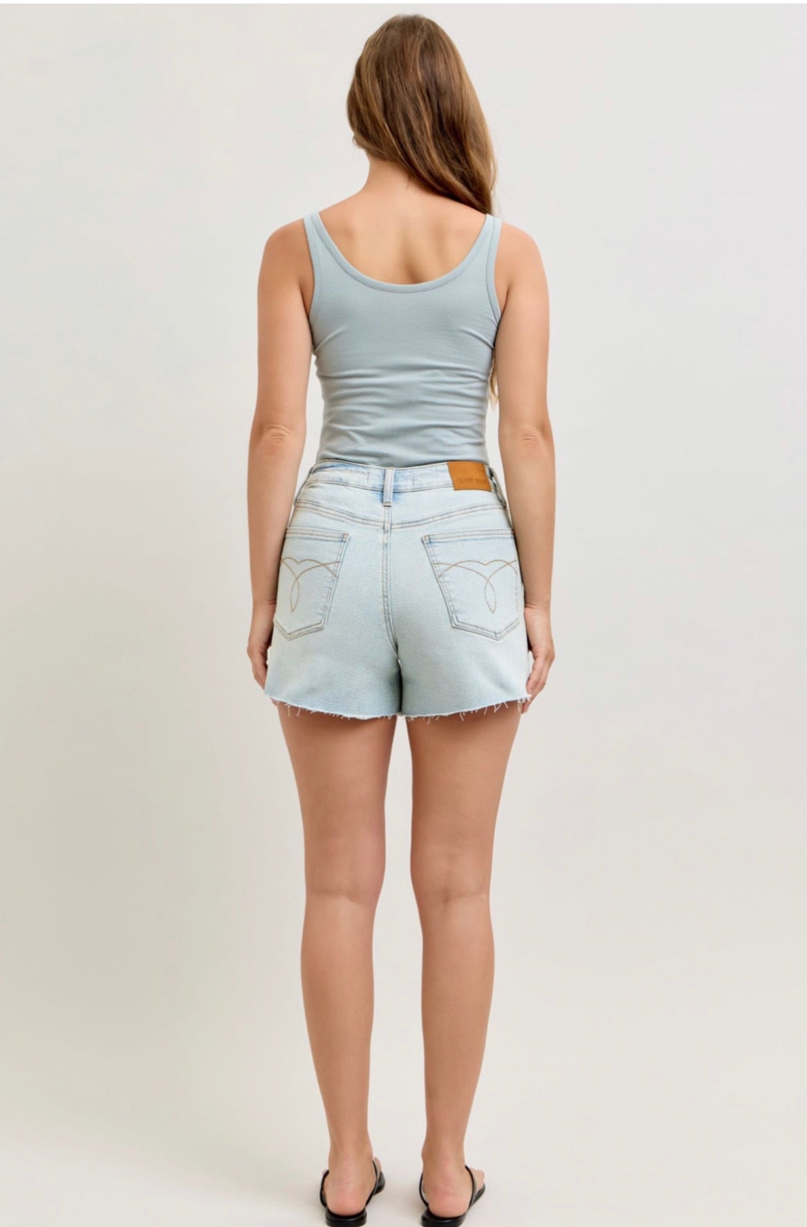Woman wearing a light blue tank top and Judy blue denim shorts on a plain background