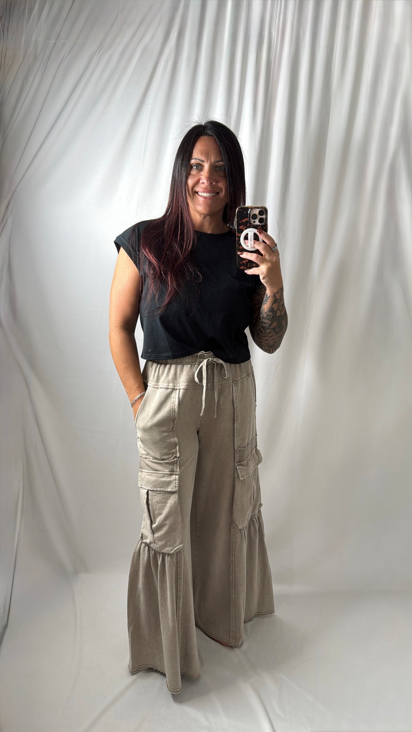A woman takes a mirror selfie wearing a black short-sleeve t-shirt and beige wide-leg cargo pants with a drawstring waist, set against a white draped fabric backdrop.