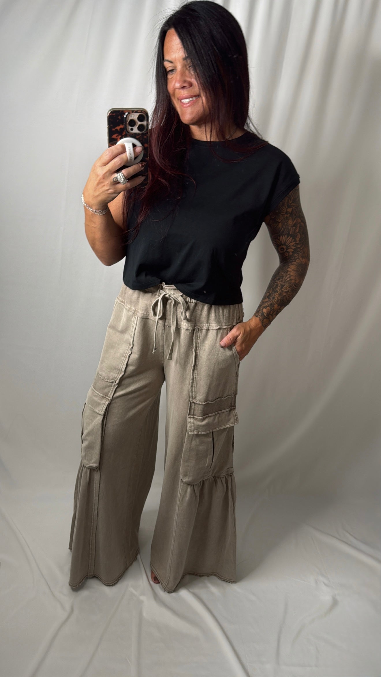 A woman takes a mirror selfie wearing a black short-sleeve t-shirt and beige wide-leg cargo pants with a drawstring waist, set against a draped white fabric backdrop.
