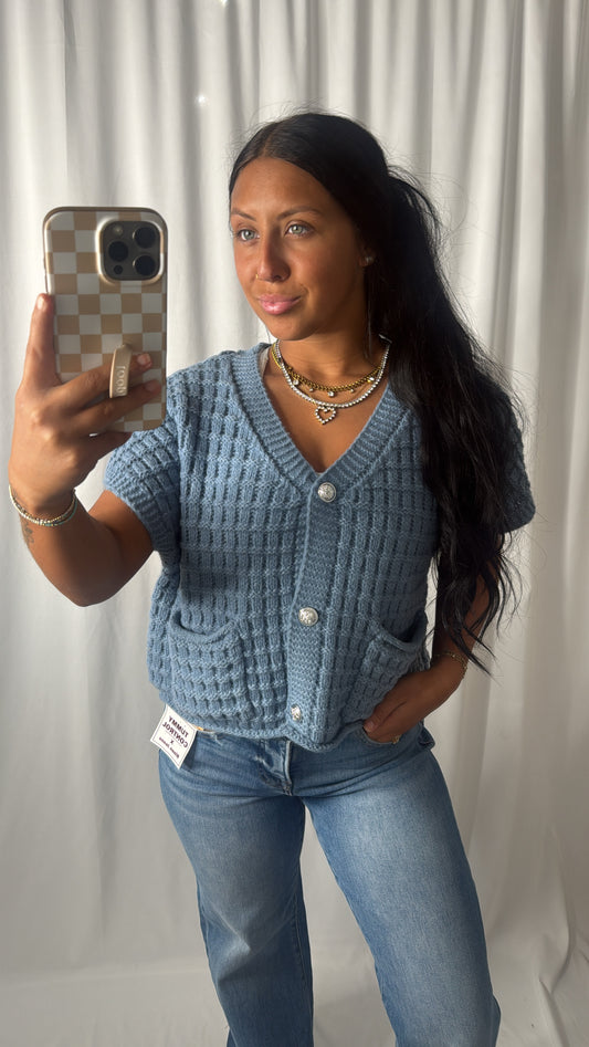 A woman takes a mirror selfie wearing a light blue textured knit cardigan with short sleeves, a V-neck, and front pockets, paired with blue jeans. She is holding a phone with a checkered case, standing against a white curtain backdrop.