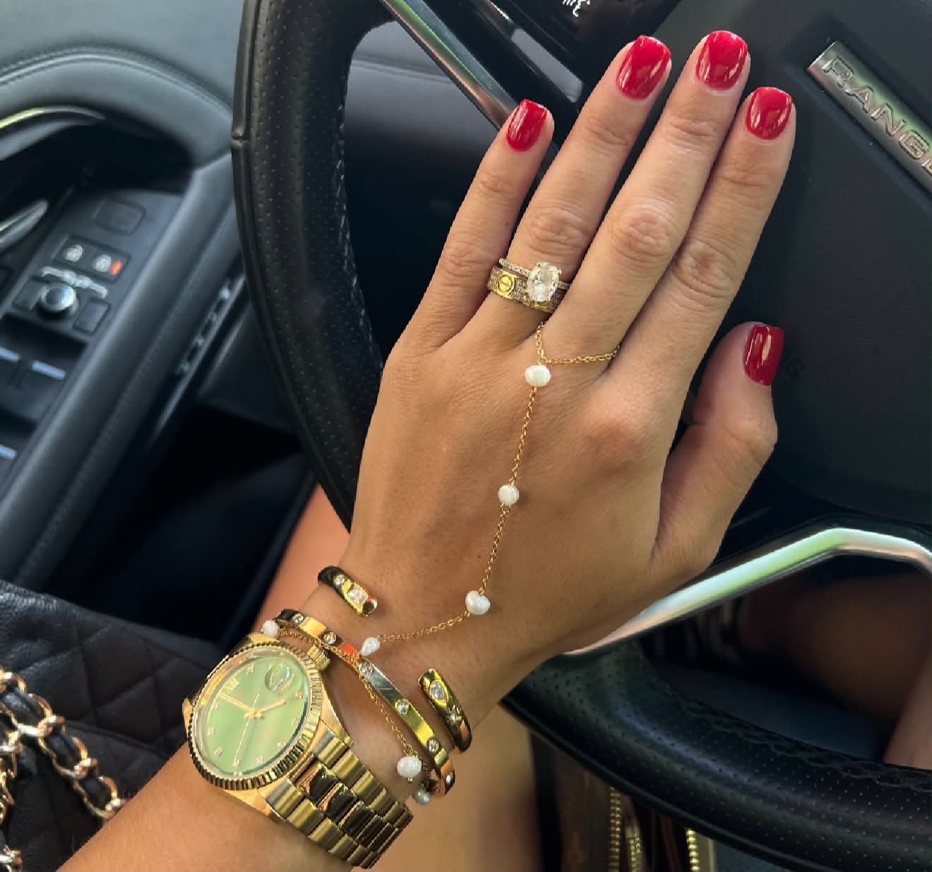 A close-up of a person's hand resting on a car steering wheel, showcasing a gold chain hand chain with white pearl beads, a gold ring, and gold bracelets. The person has bright red nail polish and is wearing a gold watch.