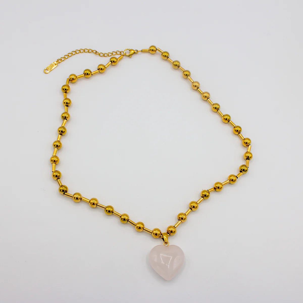 Gold beaded necklace with a heart-shaped pink stone pendant on a light gray background