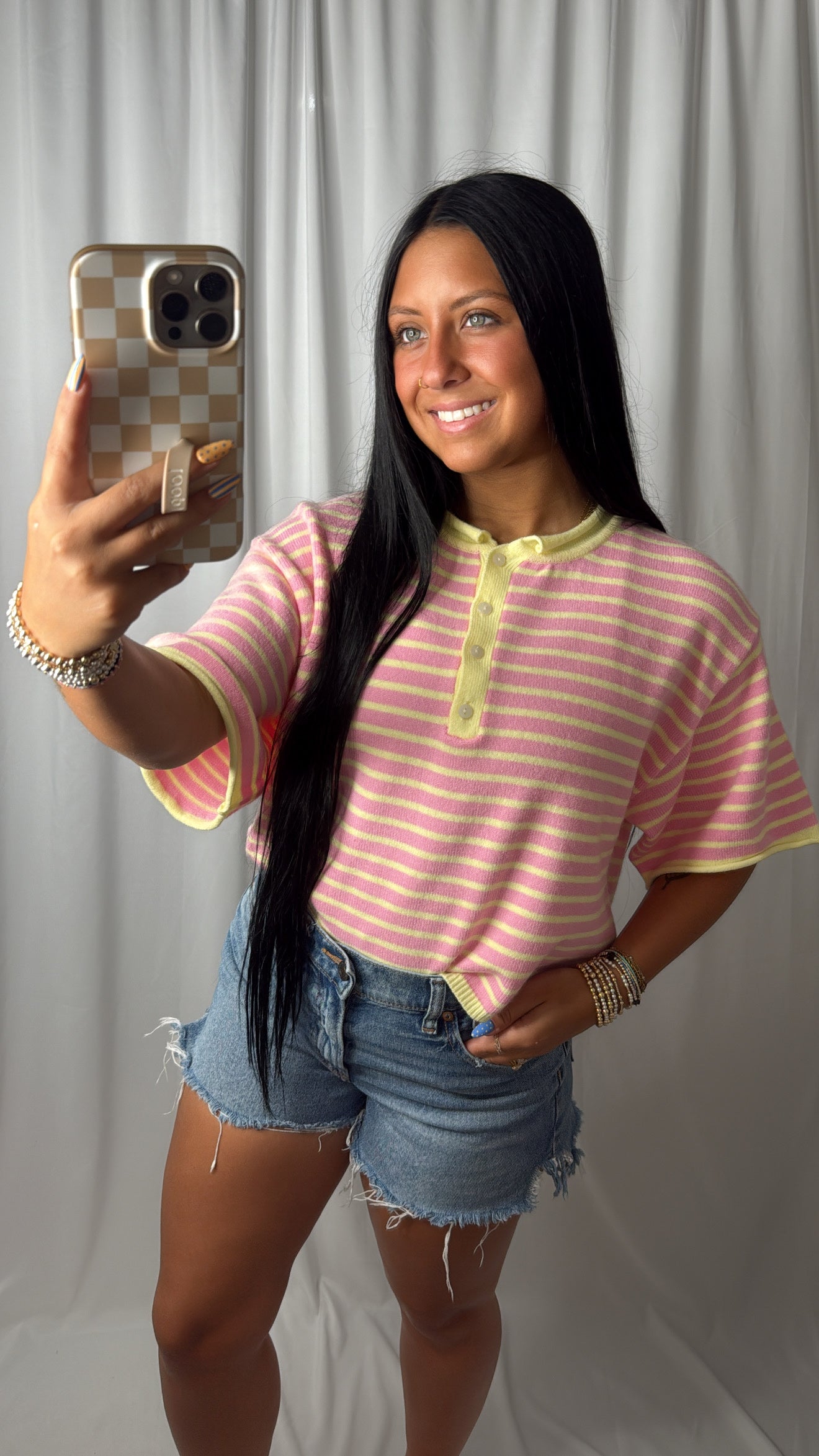 A woman taking a mirror selfie wearing a pink and white striped short-sleeve henley top with yellow trim, paired with denim shorts, against a white curtain backdrop.