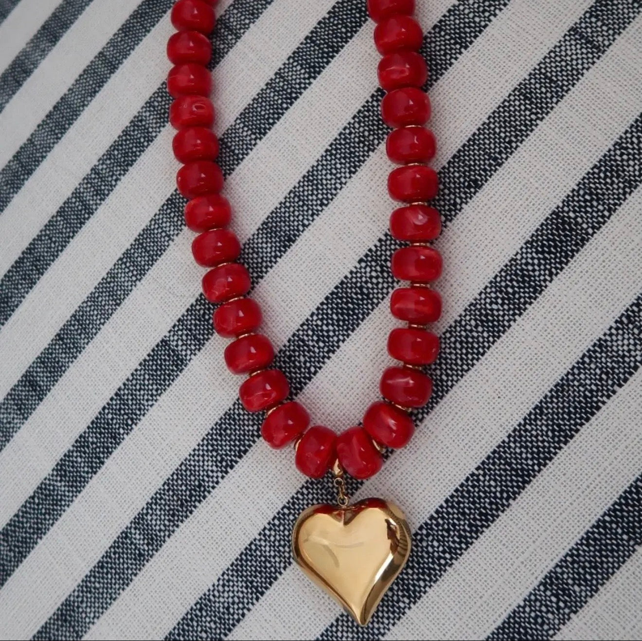 A red beaded necklace with a shiny gold heart pendant, laid flat on a black and white striped fabric background.
