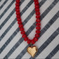 A red beaded necklace with a shiny gold heart pendant, laid flat on a black and white striped fabric background.