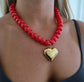 A close-up of a woman wearing a chunky red beaded necklace with a shiny gold heart-shaped pendant.