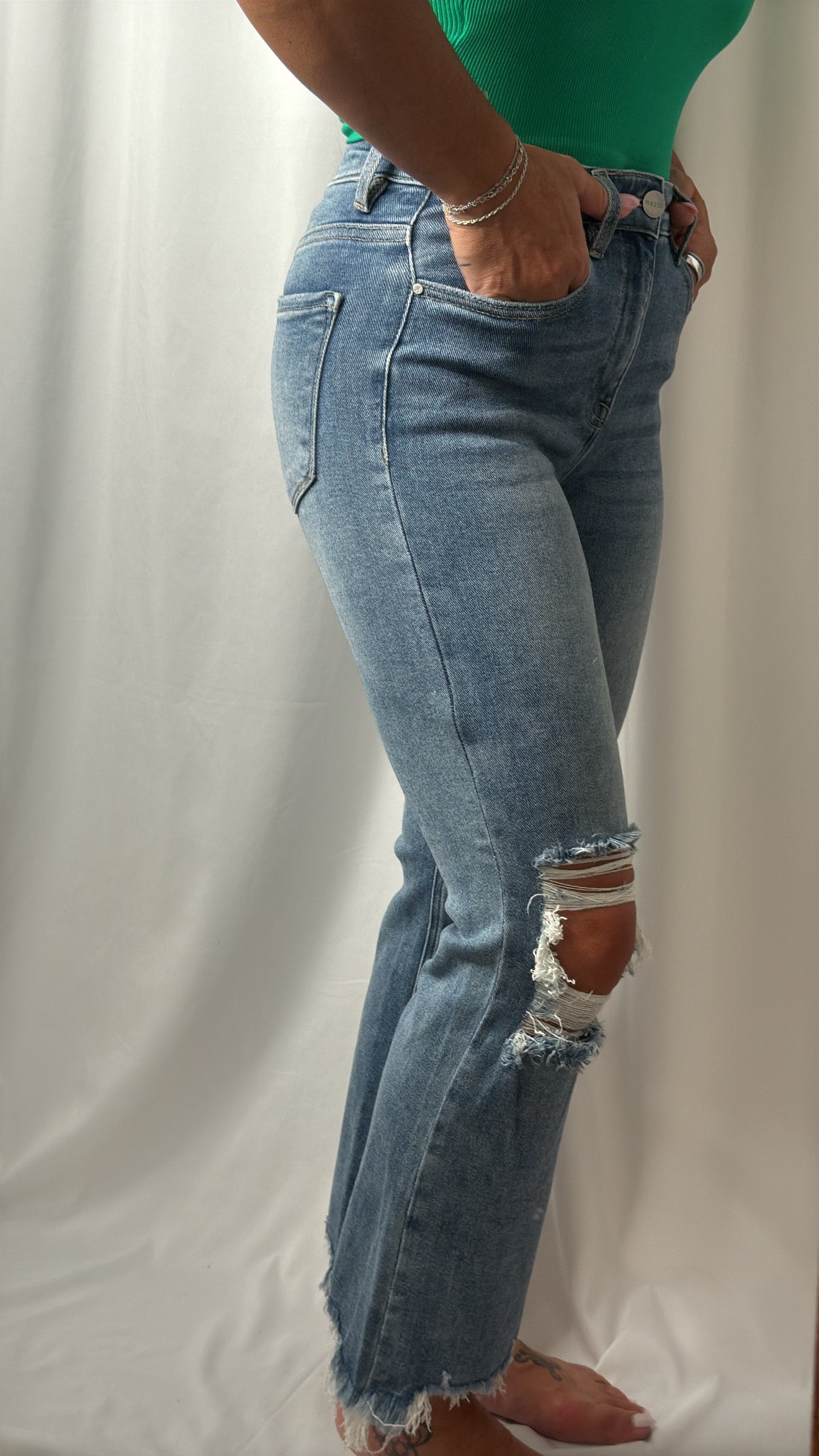A person wearing light-wash blue distressed jeans with ripped details on the knees and frayed hems, paired with a green top, standing against a white draped fabric background.