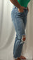 A person wearing light-wash blue distressed jeans with ripped details on the knees and frayed hems, paired with a green top, standing against a white draped fabric background.