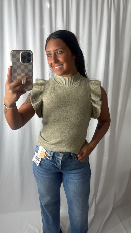 A woman takes a mirror selfie wearing a light olive green knit sweater with ruffled sleeves and blue jeans, standing in front of a white curtain backdrop.
