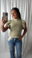 A woman takes a mirror selfie wearing a light olive green knit sweater with ruffled sleeves and blue jeans, standing in front of a white curtain backdrop.