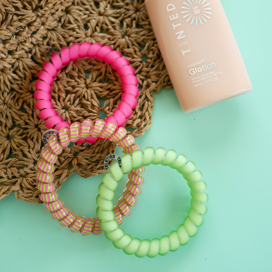 Three colorful hair ties on a textured surface with a bottle of sunscreen in the background.