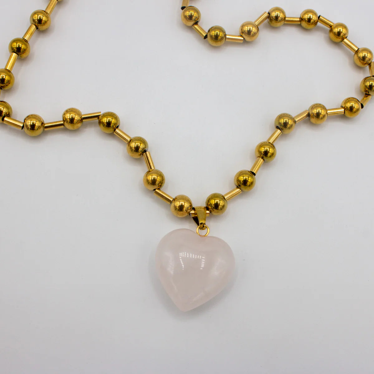 Gold beaded necklace with a heart-shaped pink pendant on a light gray background