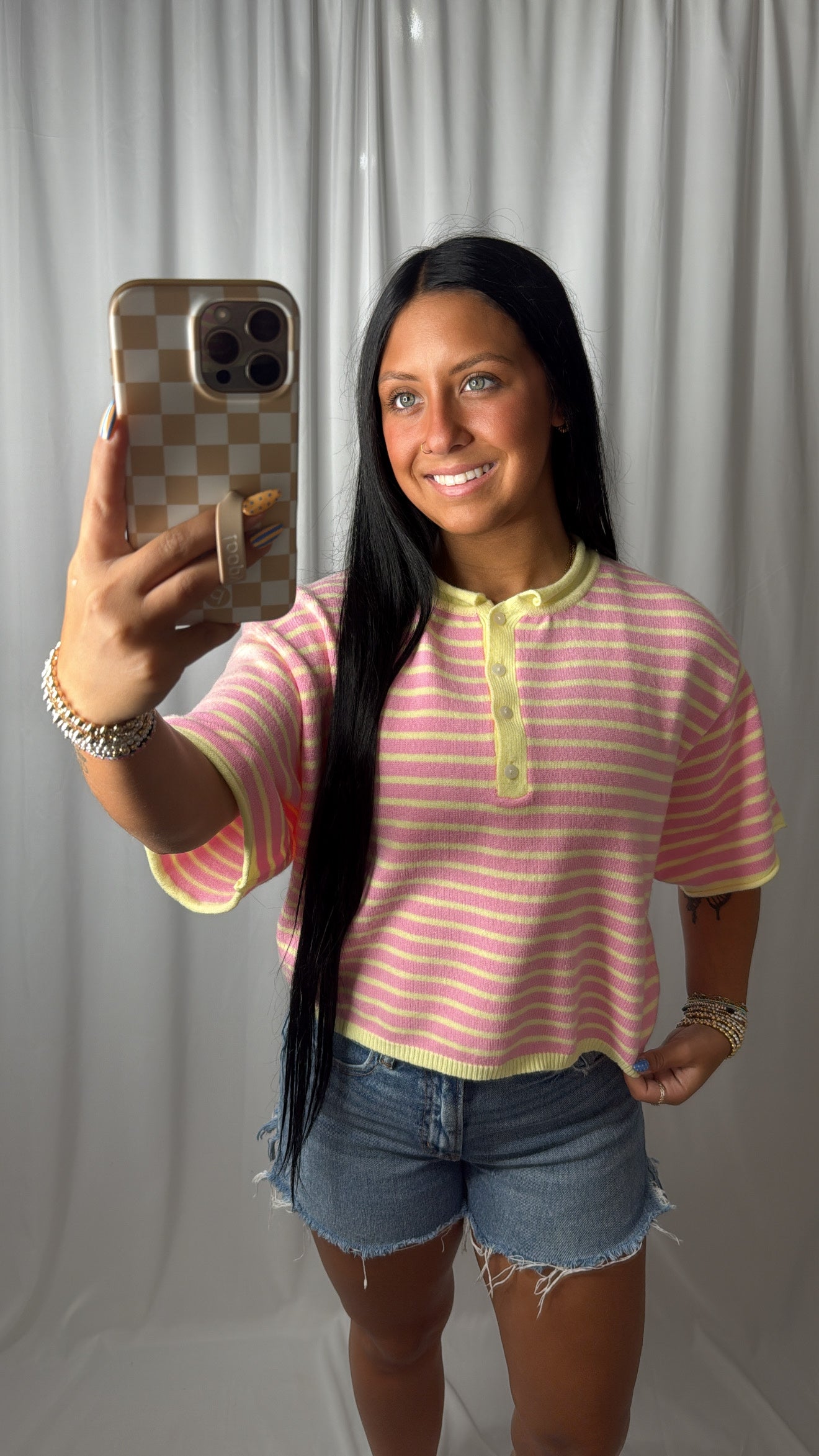 A woman takes a mirror selfie wearing a pink and white striped short-sleeve henley shirt with yellow-green trim, paired with denim shorts, in front of a white curtain backdrop.