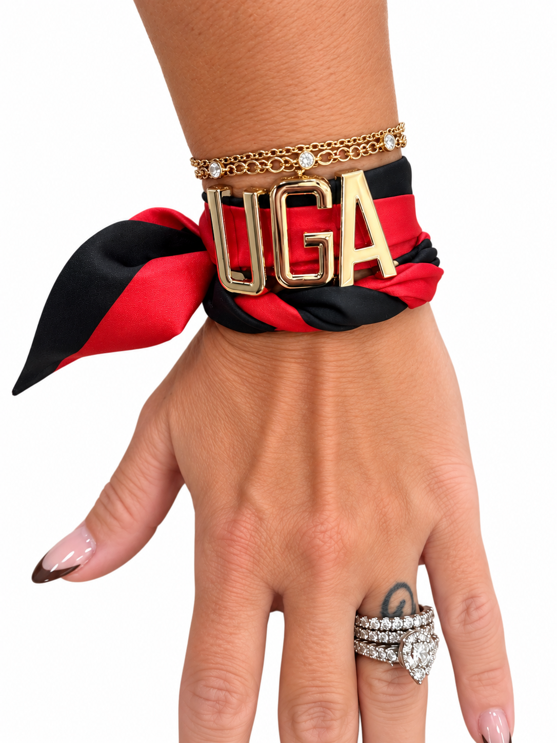 A close-up of a hand wearing a red and black satin scarf bracelet with large gold 'UGA' letter charms, layered with two gold chain bracelets, and a silver ring on the finger, against a plain white background.